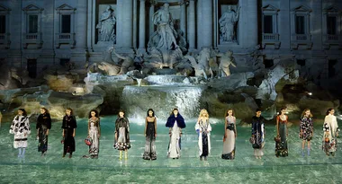 Fashion models in elegant dresses stand before Trevi Fountain, Rome, during a night runway show.