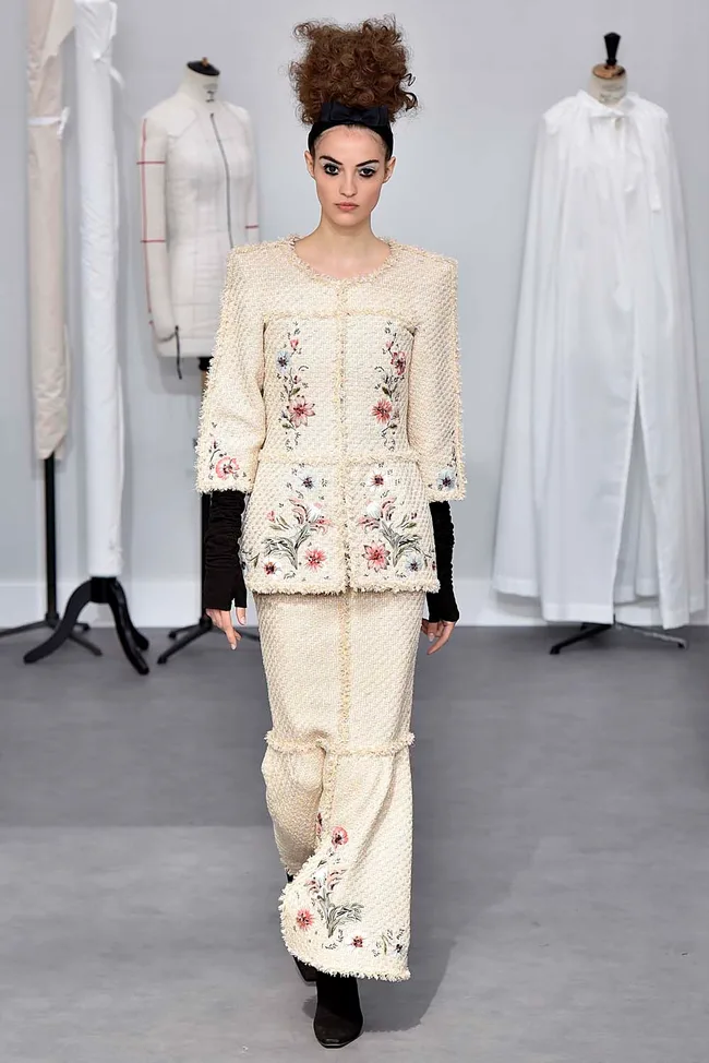 Model in cream floral embroidered outfit with high-waisted skirt on runway, mannequin and white robe in background.
