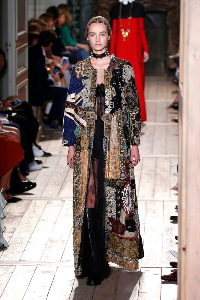 Model in an ornate, patterned gown walks on a wooden runway at a fashion show, audience seated on either side.