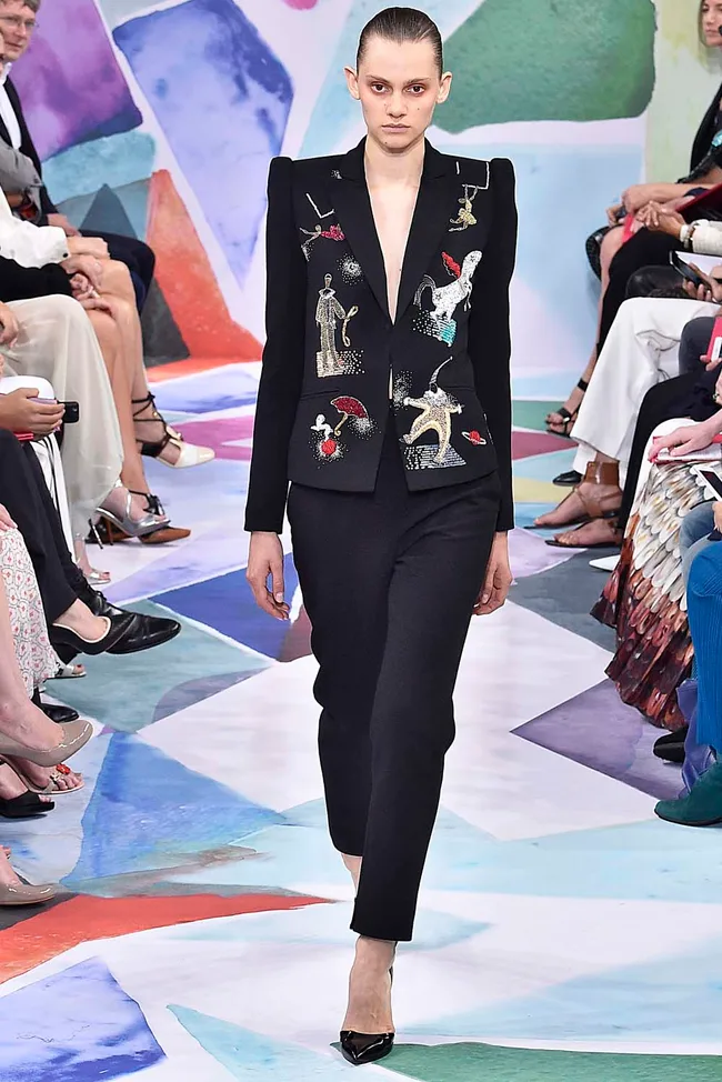 Model walking the runway in a black suit with colorful embroidered designs, on a colorful abstract background.