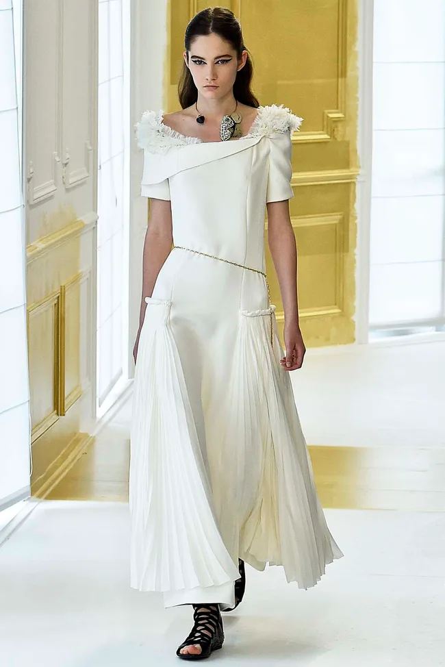 Model in an off-shoulder white dress with pleated skirt, walking on a runway with gold accents.