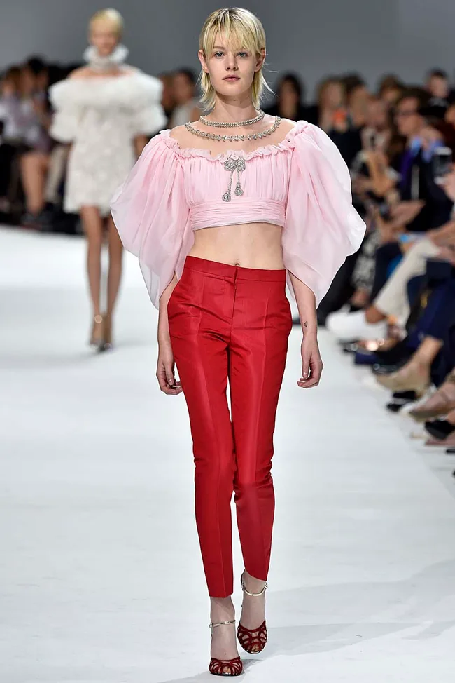 Model in a pink off-shoulder top with red pants walks on a runway with an audience in the background.