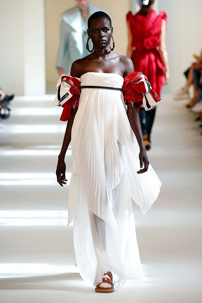Model on runway in a white pleated dress with red accents, walking confidently, with audience blurred in background.