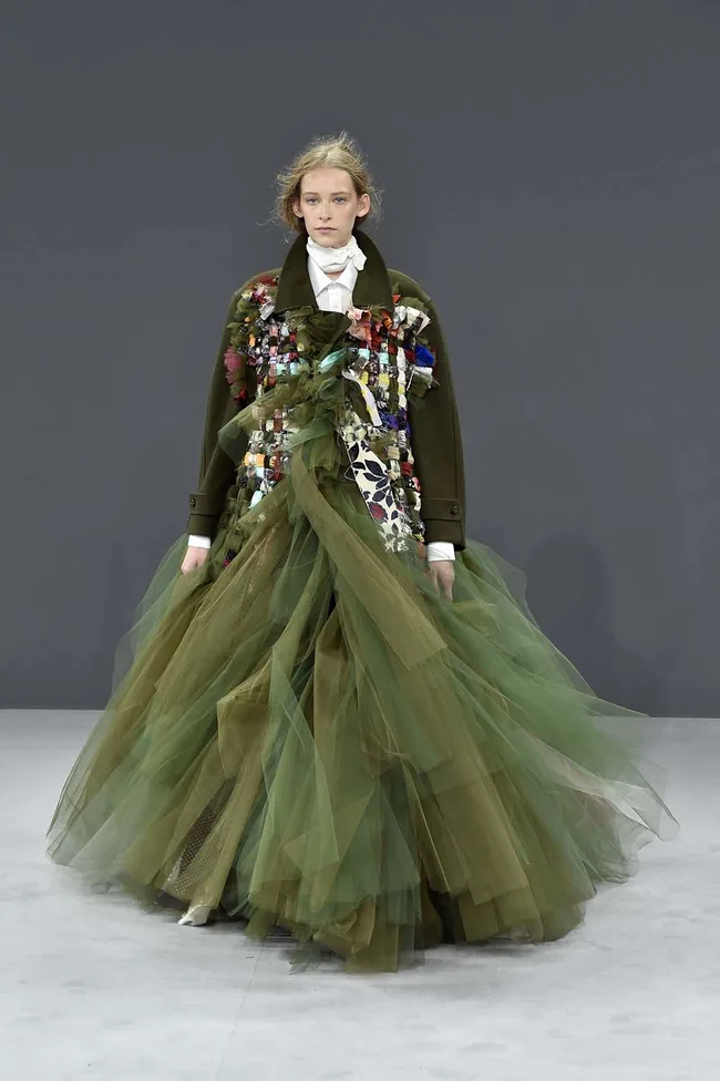 Model wearing an elaborate green tulle gown with a decorated top, walking on a gray runway.