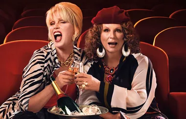 Two women sitting in theater seats, holding champagne glasses, smiling and appearing excited.