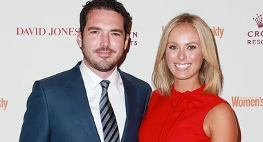 A man in a blue suit and a woman in a red dress smiling at an event, with branded backdrop logos in the background.