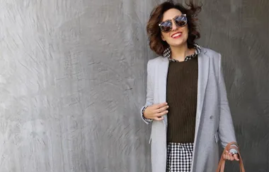 A person in sunglasses wearing a gray coat over patterned shirt and sweater, holding a brown bag, smiling.