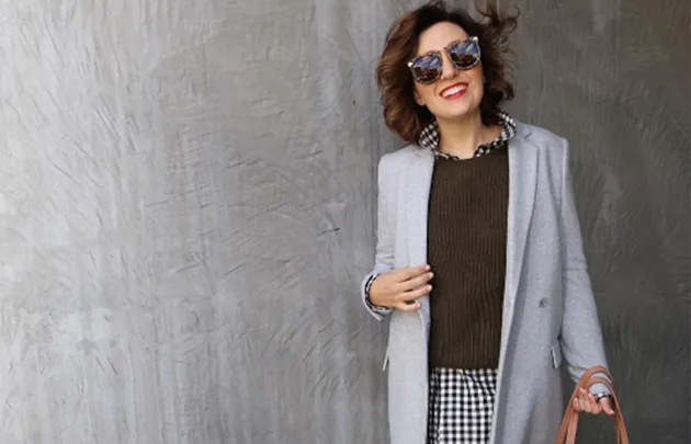 A person in sunglasses wearing a gray coat over patterned shirt and sweater, holding a brown bag, smiling.