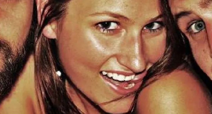 Young woman smiling with two people partially visible in the background, close-up shot.