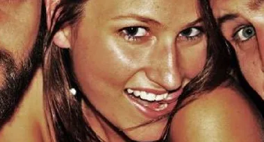 Young woman smiling with two people partially visible in the background, close-up shot.