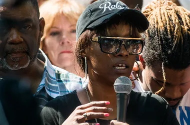 Group of people gather at an outdoor event, a woman in a cap and sunglasses speaks into a microphone.