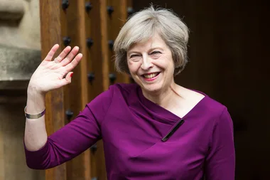 Woman with short hair in a purple top smiling and waving in front of a wooden door.