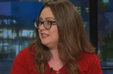 A woman with long hair and glasses, wearing a red top, is speaking in a dimly lit studio.