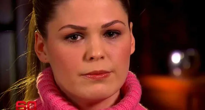 A woman with light brown hair tied back, wearing a pink turtleneck sweater, appears serious in a close-up shot.