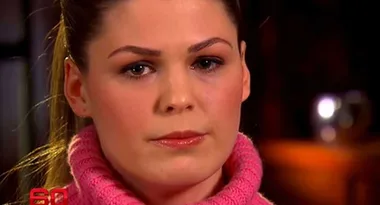 A woman with light brown hair tied back, wearing a pink turtleneck sweater, appears serious in a close-up shot.