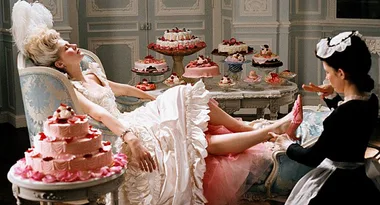 A woman in an elaborate gown lounges with desserts around her while a maid attends to her feet in a lavish room.