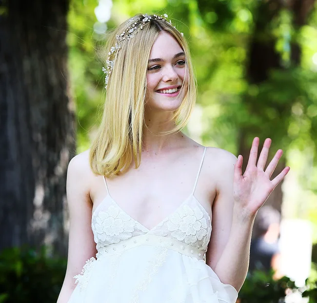 A woman with blonde hair wearing a floral white dress and a jeweled headband, smiling and waving in a green outdoor setting.