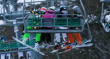 Four snowboarders in colorful attire sit on a ski lift, heading up a snowy slope.