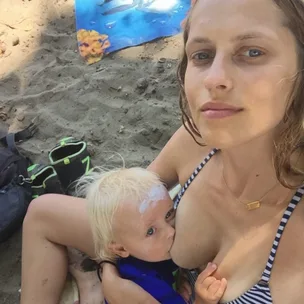 Woman in a striped swimsuit breastfeeding a baby on a sandy beach.