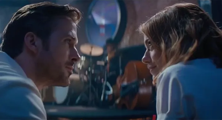 A man and a woman gaze at each other closely in a dimly lit room with musical instruments in the background.