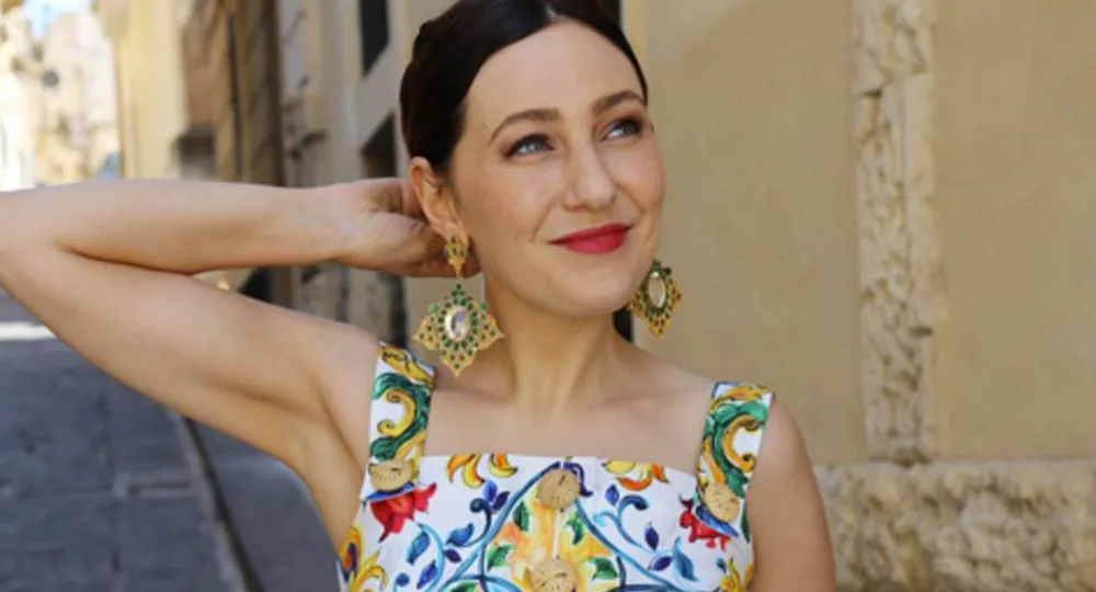A woman with dark hair smiles, wearing a colorful dress and large earrings, in a sunny outdoor setting.