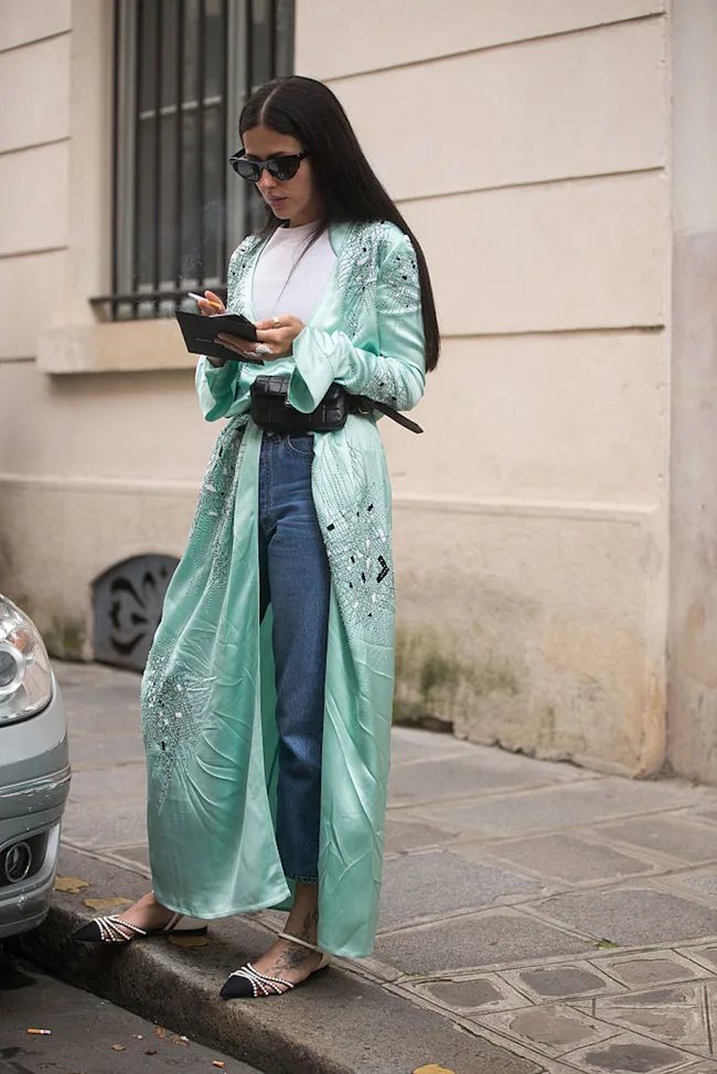 Woman wearing a long mint kimono over jeans and a white top, standing on a sidewalk, writing in a notebook.