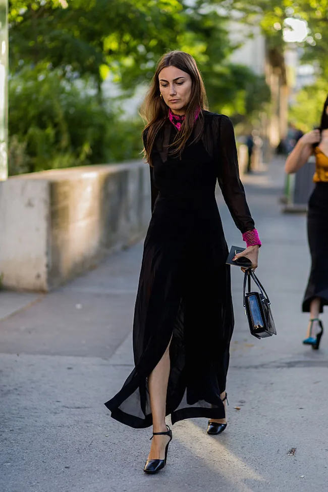 Woman in a flowing black dress walks on a sidewalk, carrying a handbag, with trees in the background.