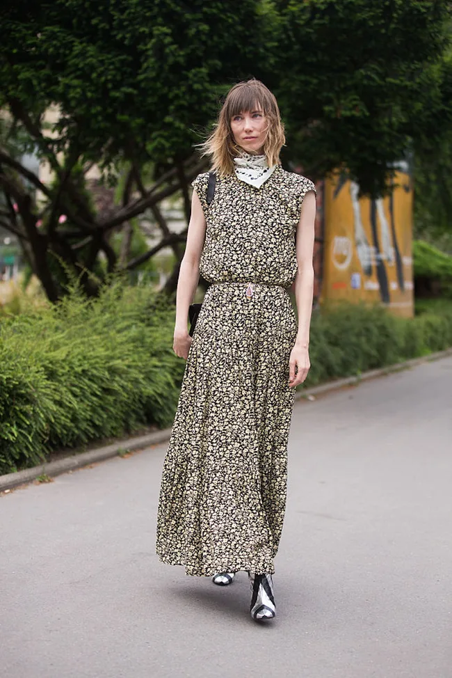 Woman in a floral dress and metallic boots walking on a path, with trees and a billboard in the background.