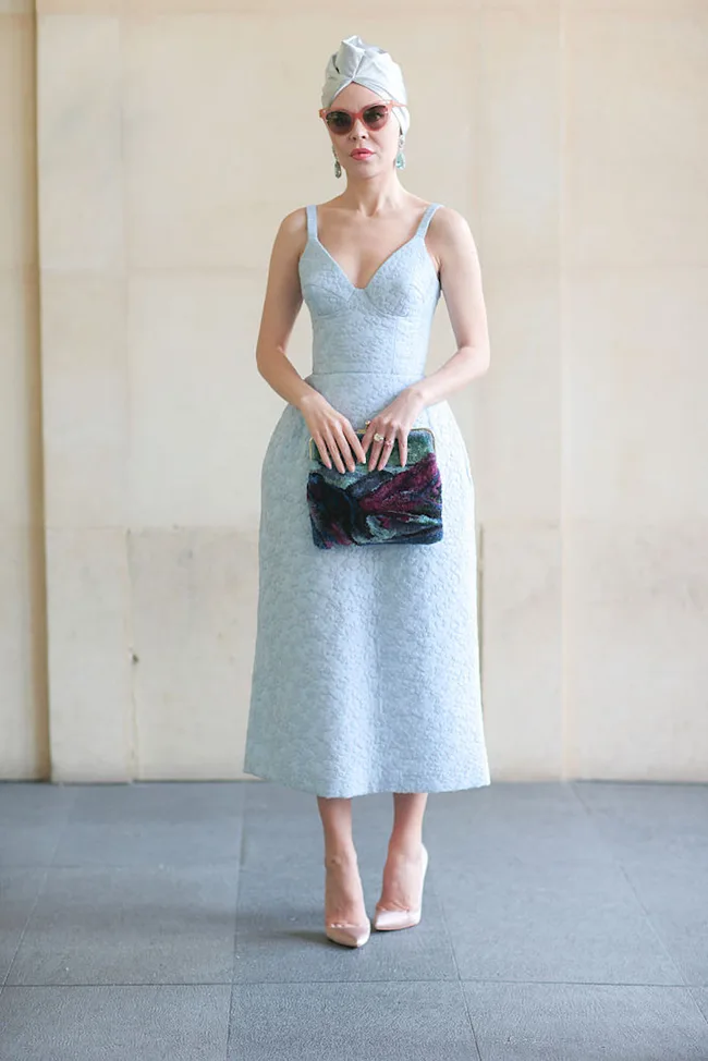 Woman in a light blue dress, sunglasses, and head wrap holding a colorful clutch, standing indoors.