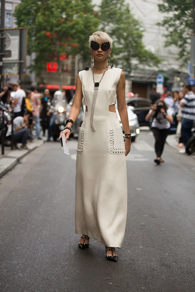 Fashionable person in a beige cutout dress, sunglasses, and black heels walking on a city street.