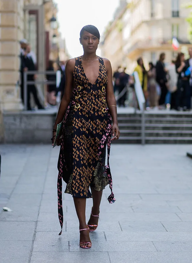 Woman in patterned dress and sandals walking on a city street, with buildings and people in the background.