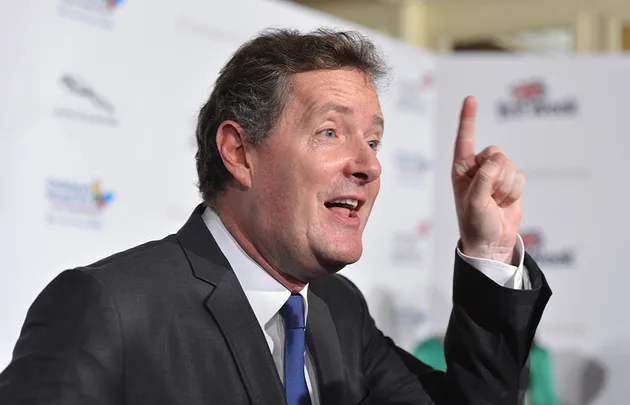 A man in a suit gestures enthusiastically with one finger raised, speaking at an event with a branded backdrop.