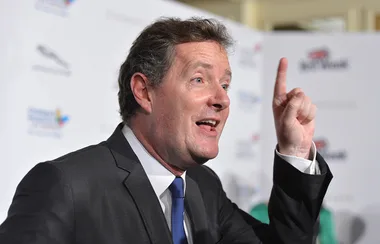 A man in a suit gestures enthusiastically with one finger raised, speaking at an event with a branded backdrop.