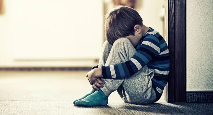 Child sitting on the floor with head down, wearing a striped shirt and gray pants, leaning against a wall.