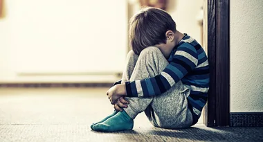 Child sitting on the floor with head down, wearing a striped shirt and gray pants, leaning against a wall.