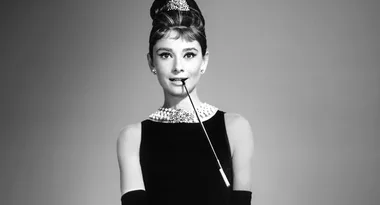 A woman in a black dress and pearl necklace holds a long cigarette holder, posing elegantly against a gray background.