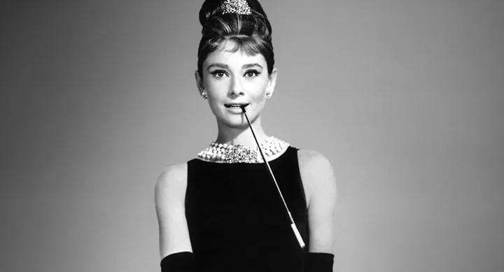 A woman in a black dress and pearl necklace holds a long cigarette holder, posing elegantly against a gray background.