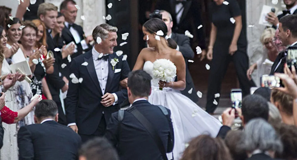 A bride and groom smile at each other as they walk through a crowd of guests throwing flower petals.