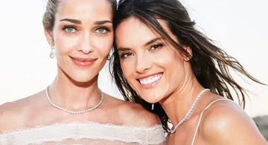 Two women smiling at the camera, wearing elegant dresses and necklaces, standing close together outdoors.