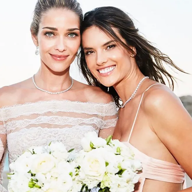 Two women smiling, holding white flower bouquets, one in a lace wedding dress, the other in a light pink dress.
