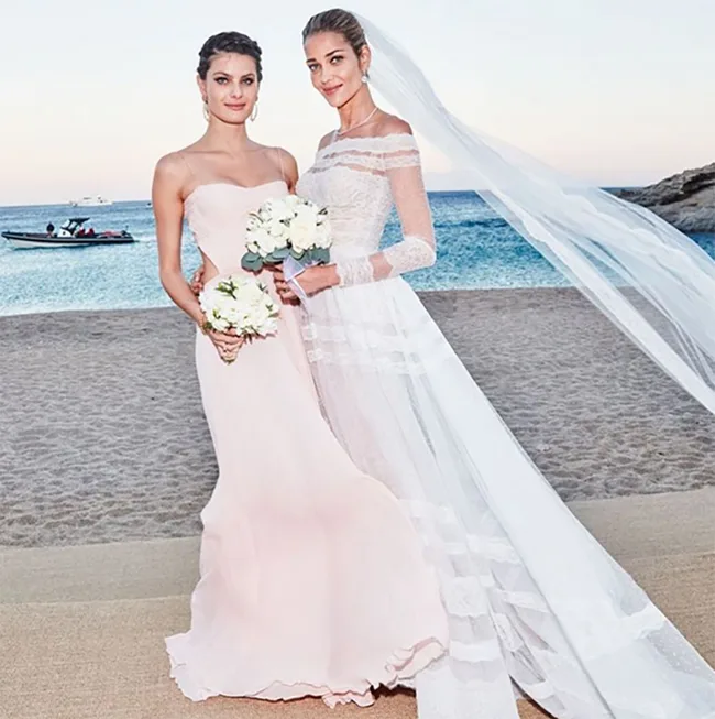 Two women, one in a white wedding gown and veil, the other in a pink dress, holding bouquets on a beach.