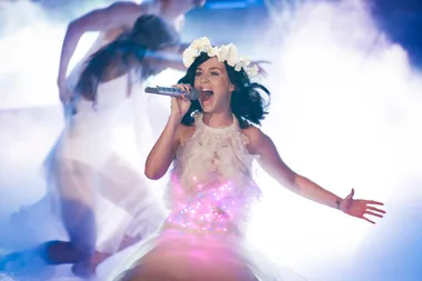 Singer in a white dress with flowers in hair performs passionately on stage with vibrant lights.