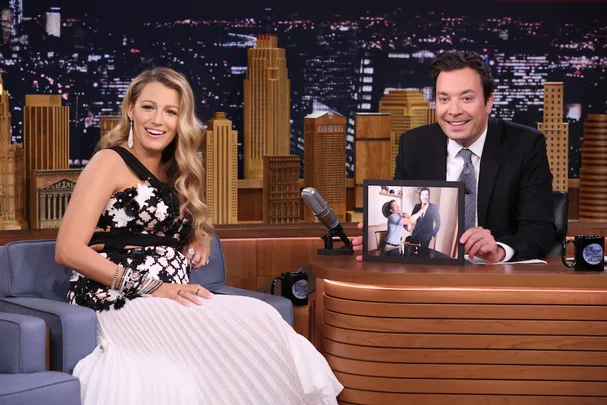 Two people sit on a talk show set; one holds a photo of suit fitting; a cityscape backdrop is visible.