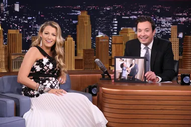 Two people sit on a talk show set; one holds a photo of suit fitting; a cityscape backdrop is visible.