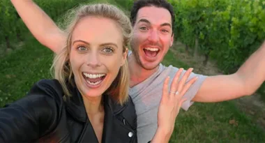 Two people smiling widely, taking a selfie in a lush green vineyard setting.
