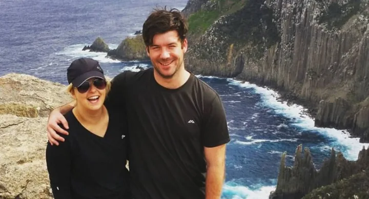A smiling couple poses with arms around each other on a cliffside, with the ocean and rocky cliffs in the background.