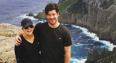 A smiling couple poses with arms around each other on a cliffside, with the ocean and rocky cliffs in the background.