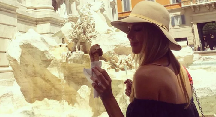 A woman in a sun hat holds an ice cream cone by a fountain in a European square.