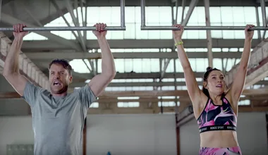 Two people hanging from monkey bars in a gym, one in a gray shirt and another in floral workout attire.