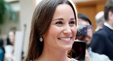 A woman with long brown hair smiling, wearing dangling earrings, in a crowded indoor setting.